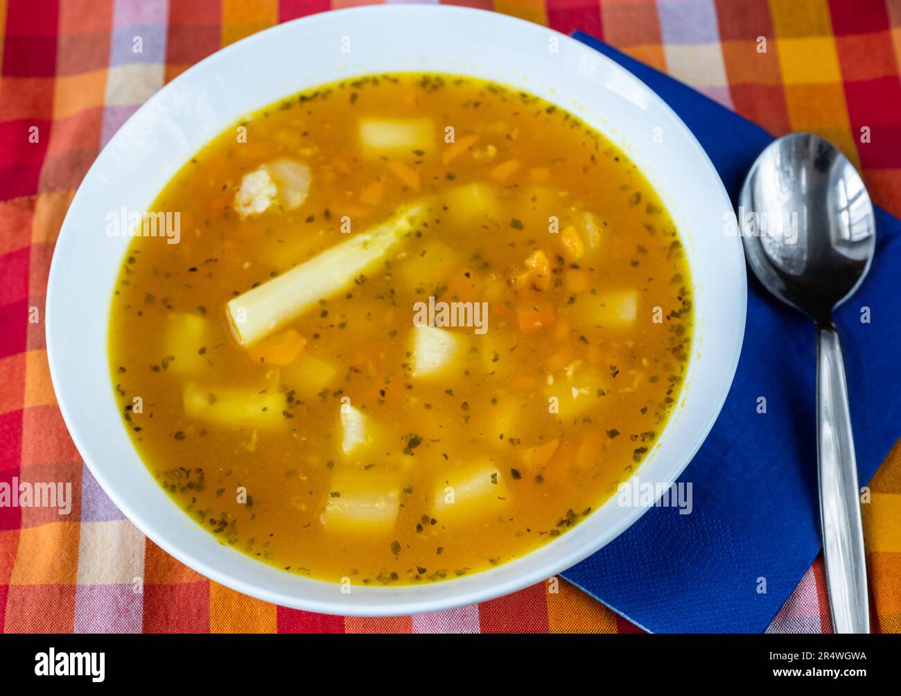 Vegetable soup with asparagus, potato, celery and carrot in white plate, spoon,blue napkin and colorful checkered tablecloth, closeup. Stock Photo