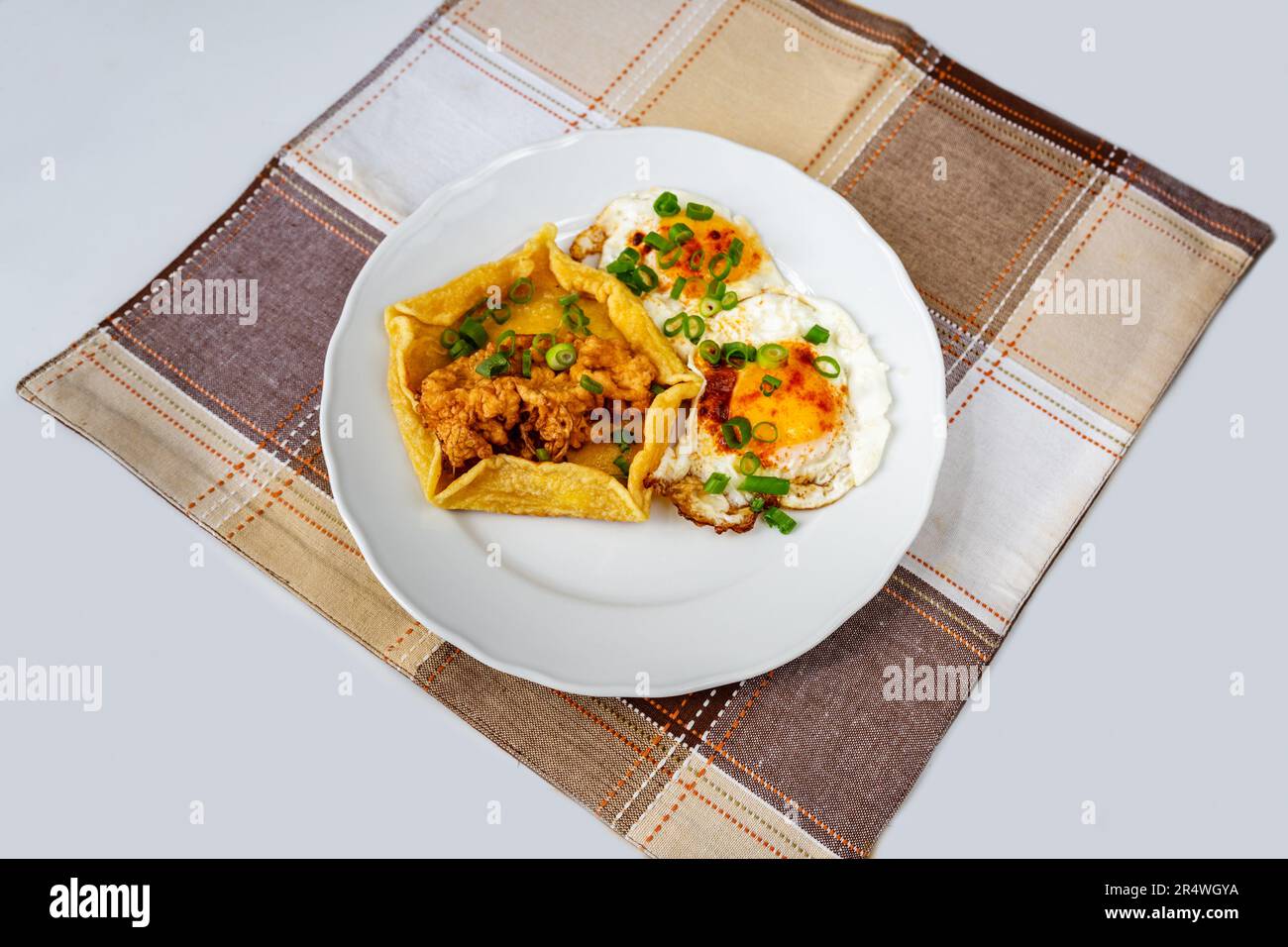 Fried egg and salty pancake with fried elderflower on plate on checkered tablecloth on white background. Stock Photo