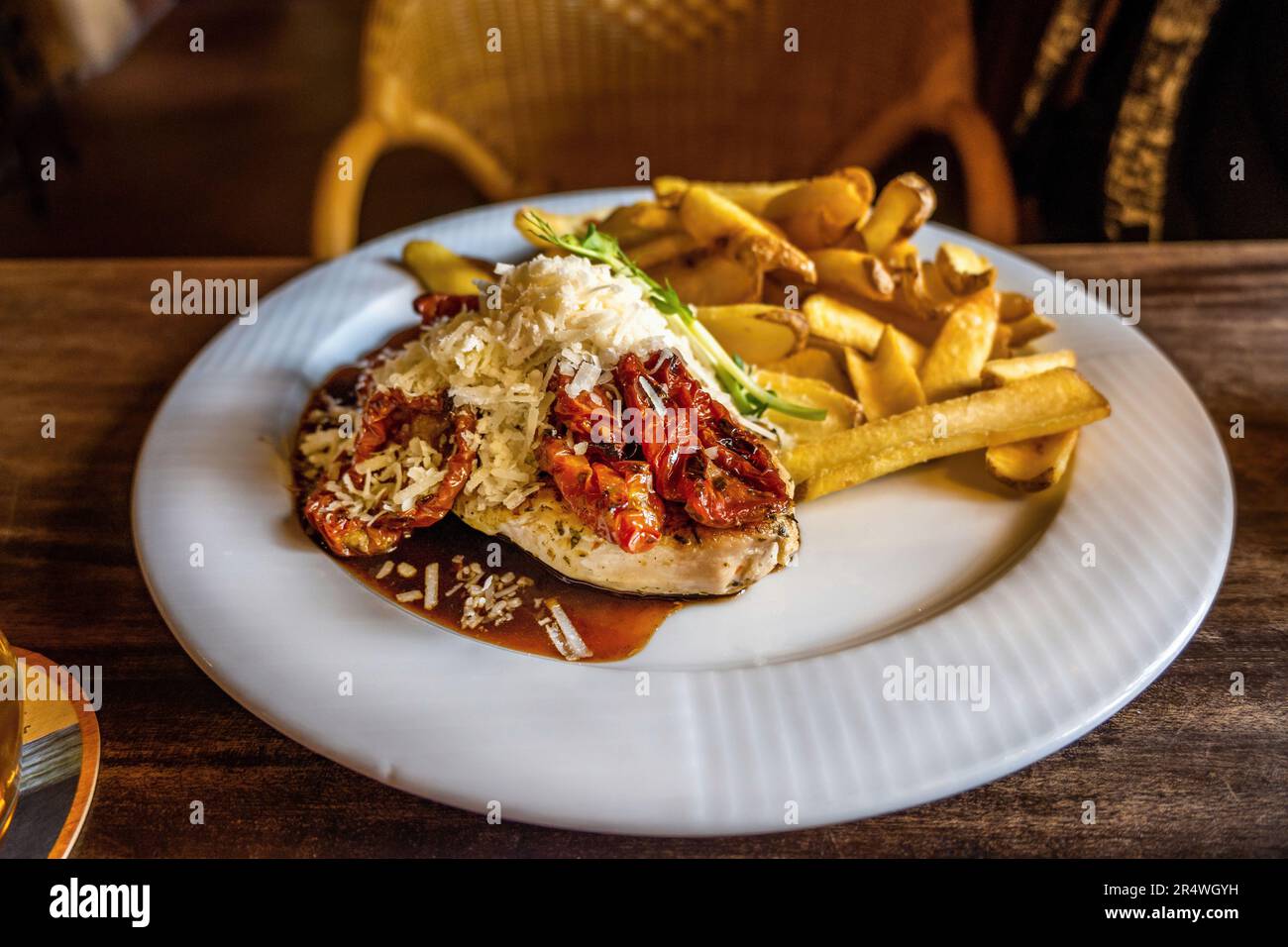 Chicken fillet with dried tomato and grated cheese with french fries on white plate, at restaurant table, closeup. Stock Photo