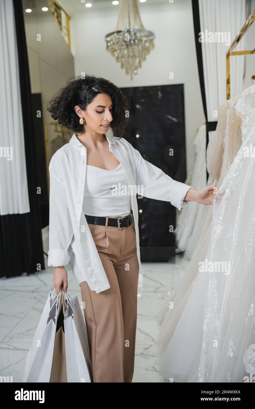 charming middle eastern woman with brunette and wavy hair standing in beige pants with white shirt and holding shopping bags while choosing wedding dr Stock Photo