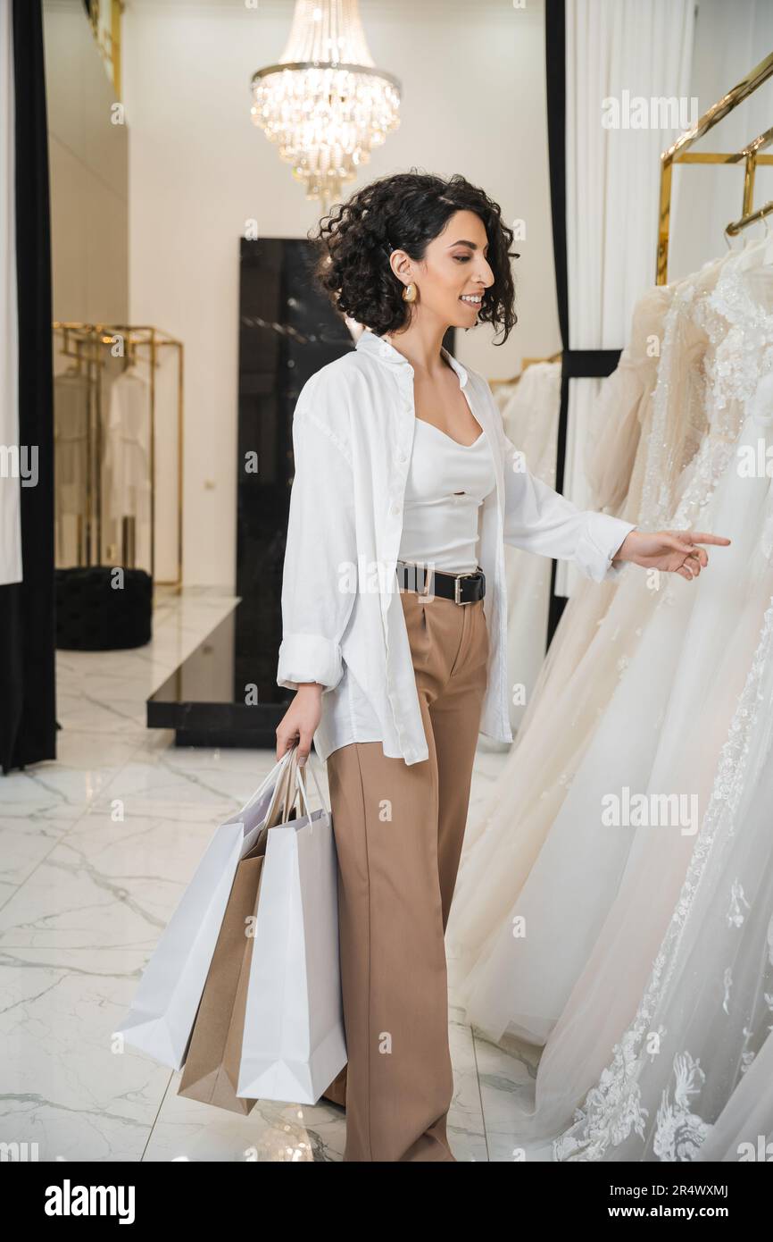stylish and happy middle eastern woman with brunette and wavy hair standing in beige pants with white shirt and holding shopping bags while choosing w Stock Photo