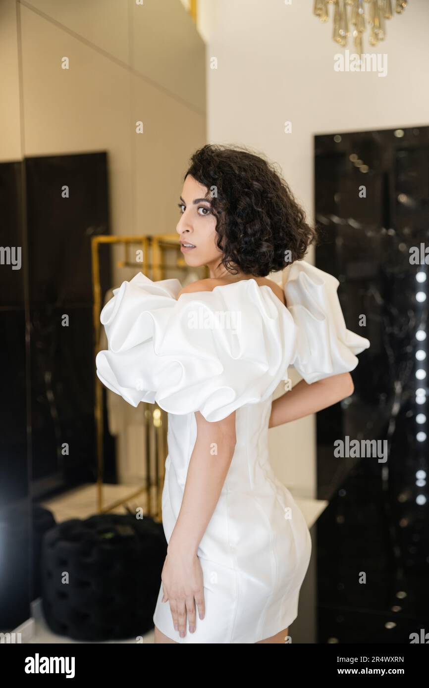 middle eastern woman with brunette wavy hair trying on trendy wedding dress with puff sleeves and ruffles and looking away in modern bridal boutique, Stock Photo