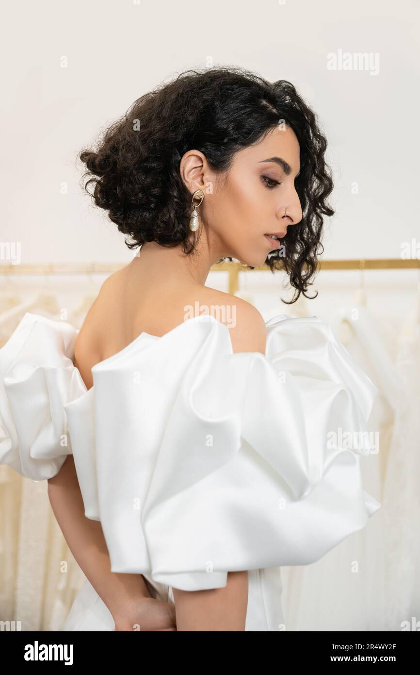 brunette middle eastern woman with brunette and wavy hair posing in trendy wedding dress with puff sleeves and ruffles in bridal boutique and looking Stock Photo