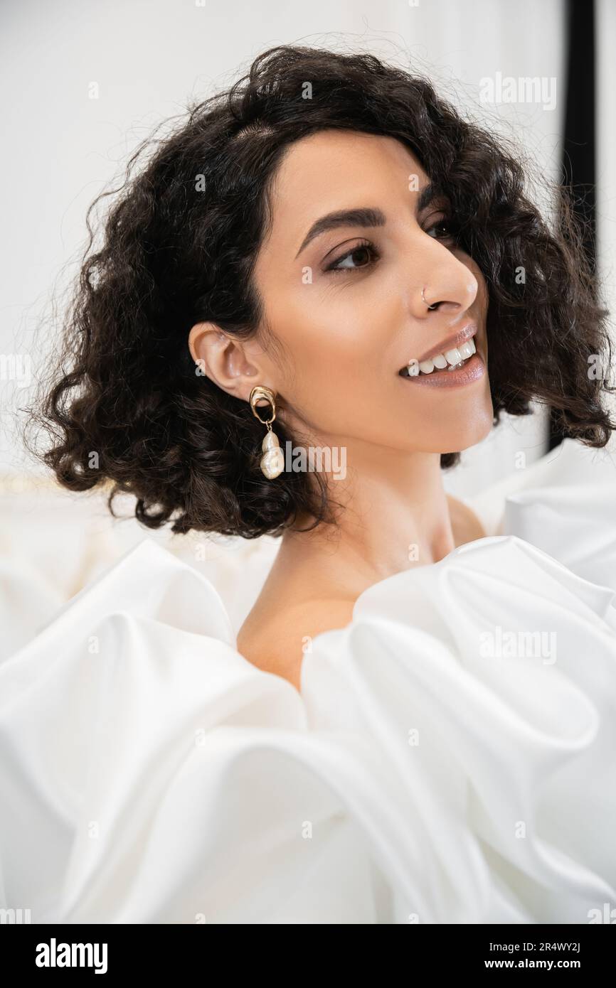 portrait of happy brunette middle eastern bride with wavy hair posing in trendy wedding dress with puff sleeves and ruffles in bridal boutique and loo Stock Photo