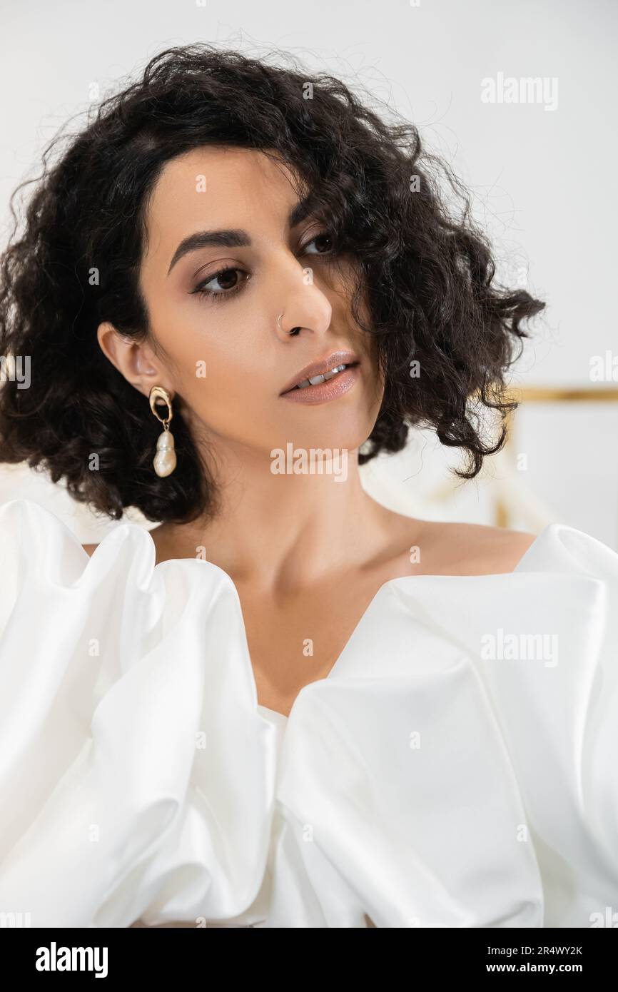 portrait of brunette middle eastern bride with wavy hair posing in trendy wedding dress with puff sleeves and ruffles in bridal boutique and looking a Stock Photo