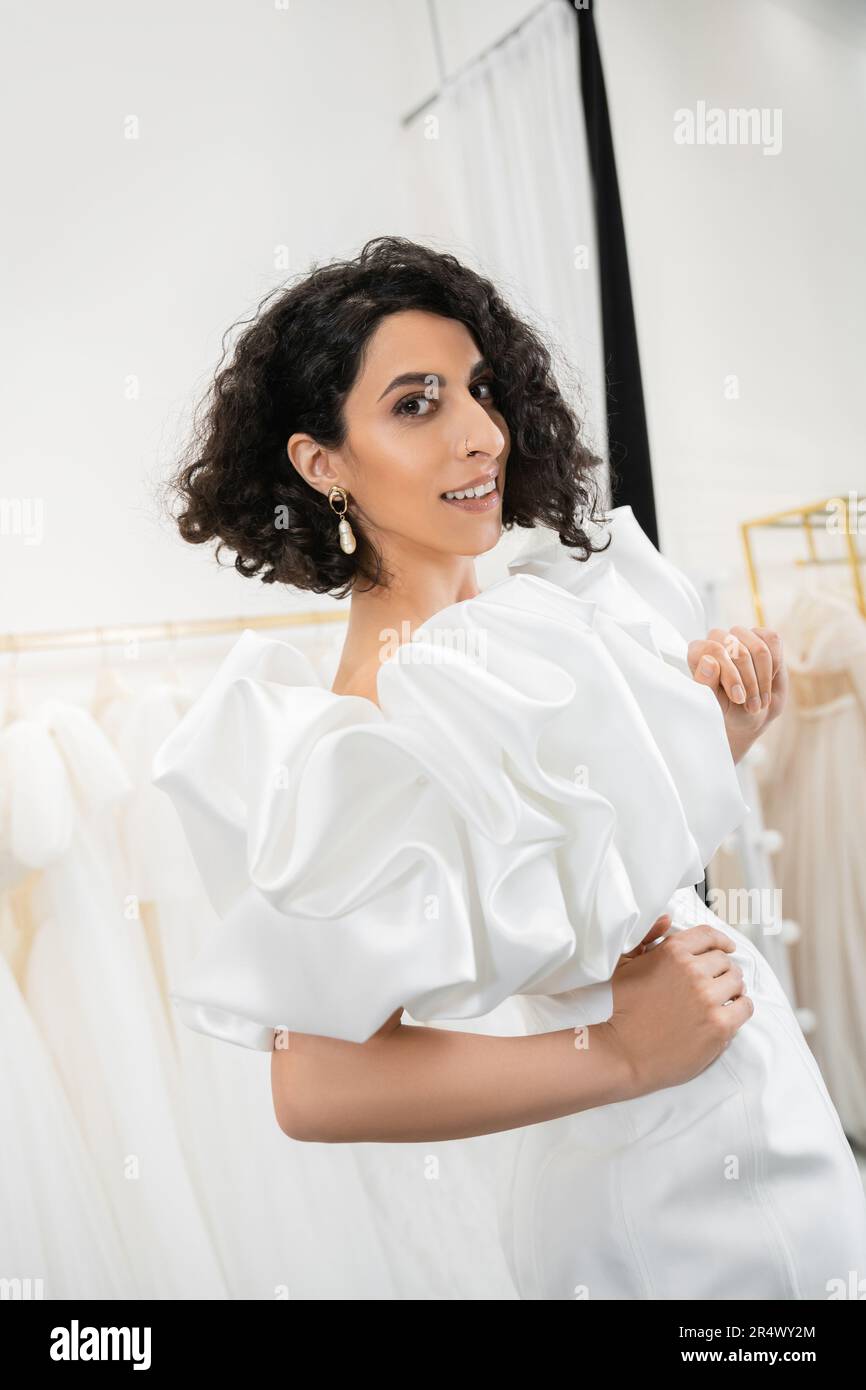 happy middle eastern bride with brunette and wavy hair posing with hands on hips in trendy wedding dress with puff sleeves and ruffles in bridal bouti Stock Photo
