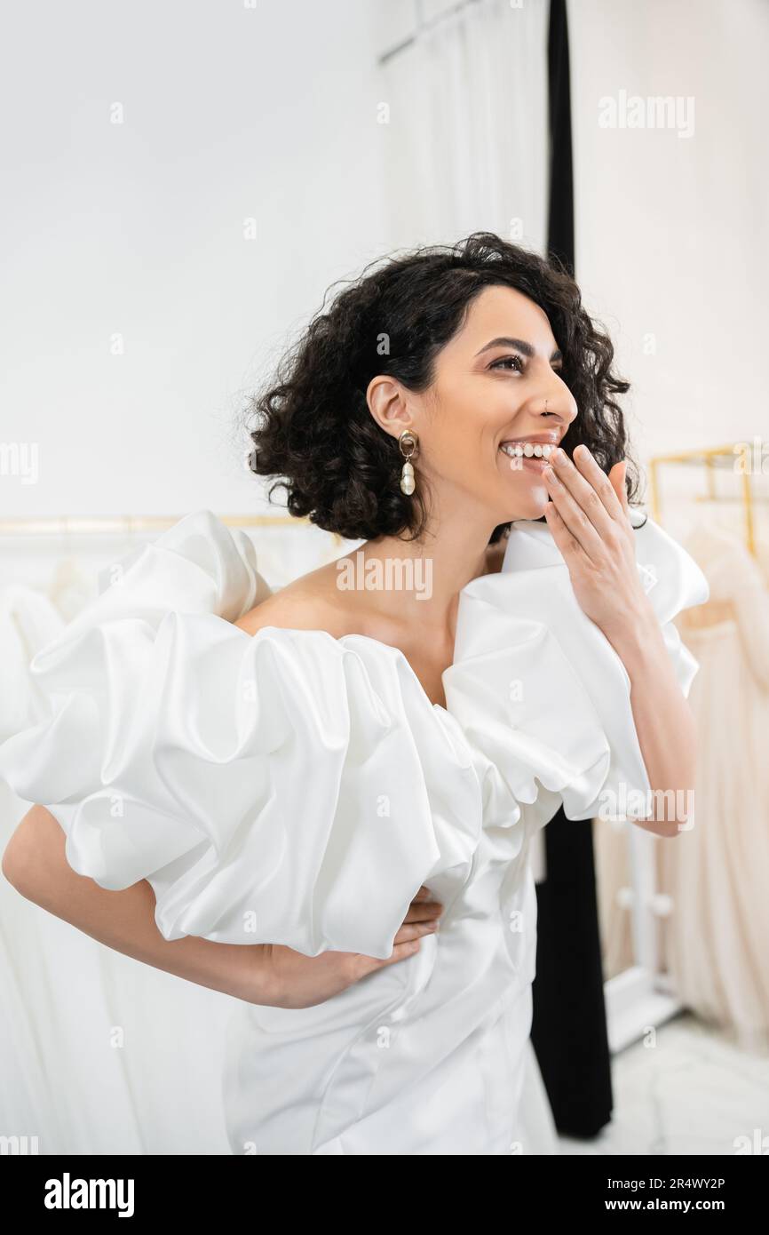 radiant middle eastern bride with brunette wavy hair laughing, covering mouth and posing in trendy wedding dress with puff sleeves and ruffles in brid Stock Photo