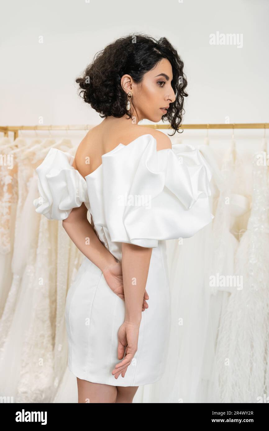 stunning middle eastern bride with brunette and wavy hair posing in stylish wedding dress with puff sleeves and ruffles in bridal boutique next to tul Stock Photo
