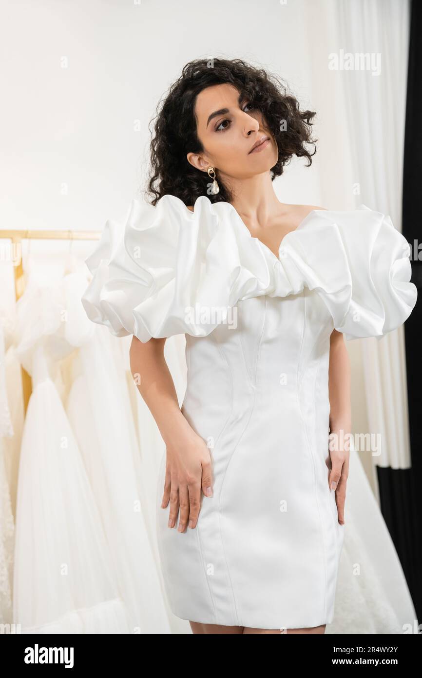 elegant middle eastern bride with brunette and wavy hair posing in trendy wedding dress with puff sleeves and ruffles in bridal boutique next to tulle Stock Photo