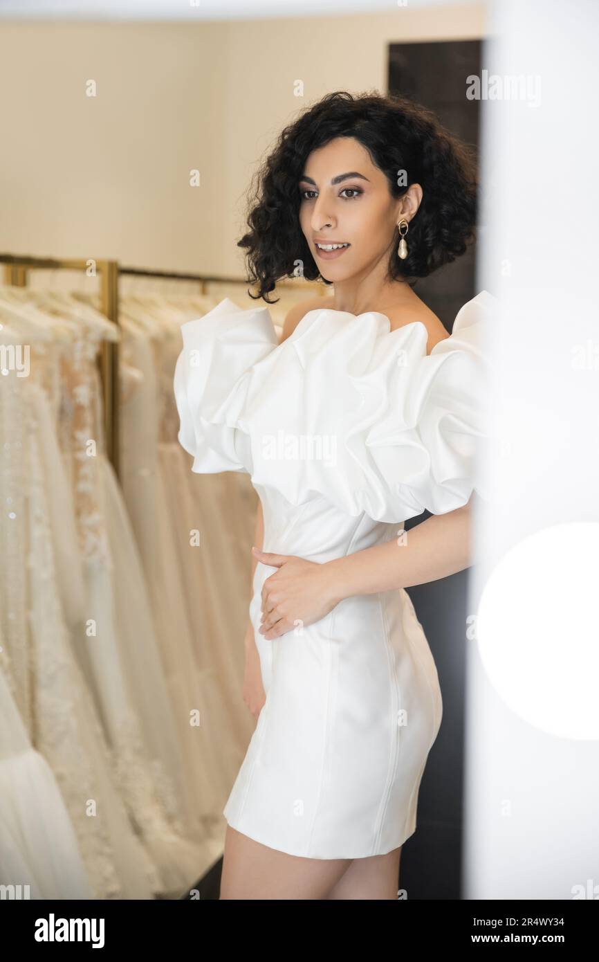 smiling middle eastern woman with brunette and wavy hair trying on trendy wedding dress with puff sleeves and ruffles near mirror in bridal boutique n Stock Photo