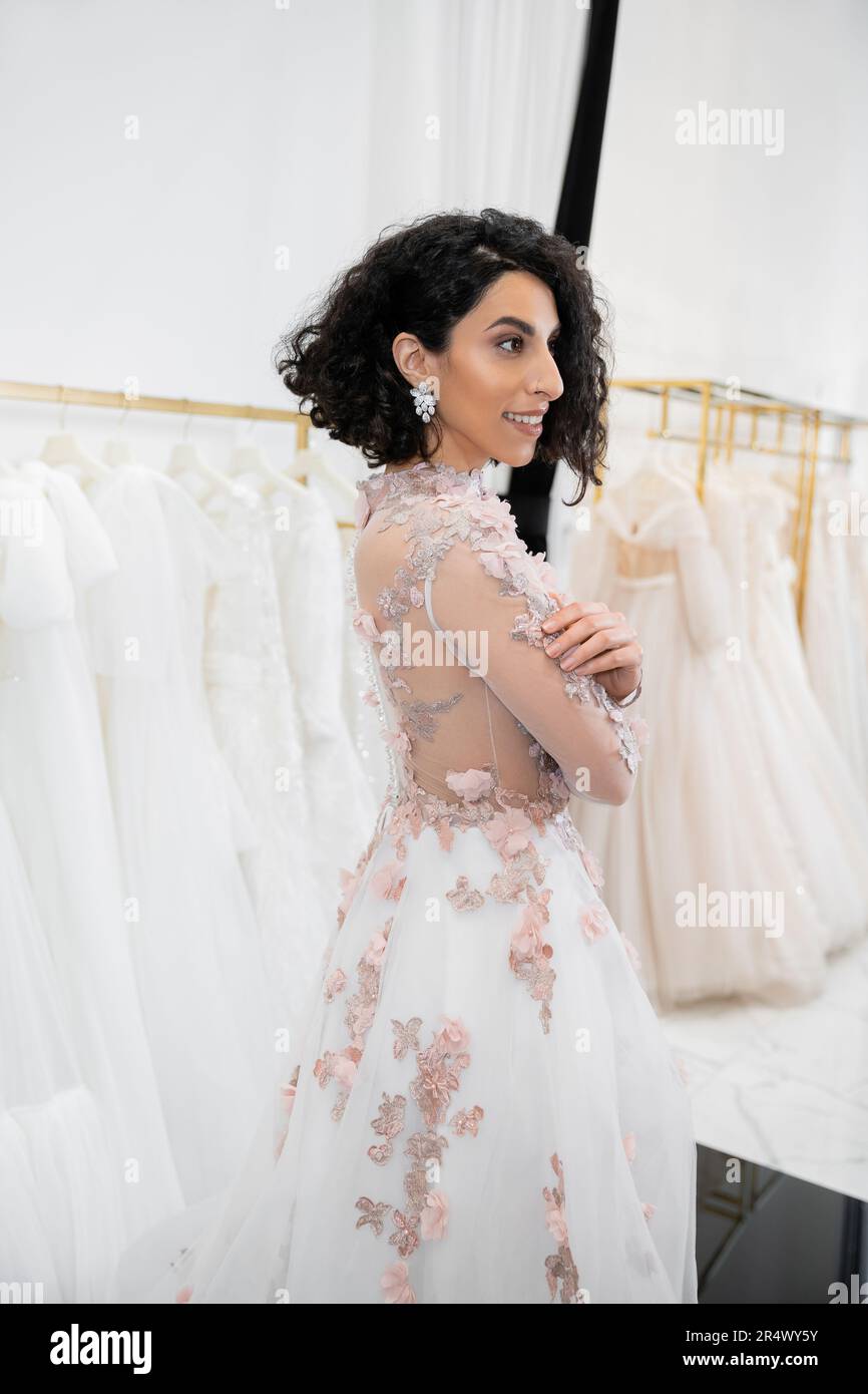 cheerful middle eastern and brunette woman with wavy hair standing in gorgeous and floral wedding dress inside of luxurious bridal salon around white Stock Photo