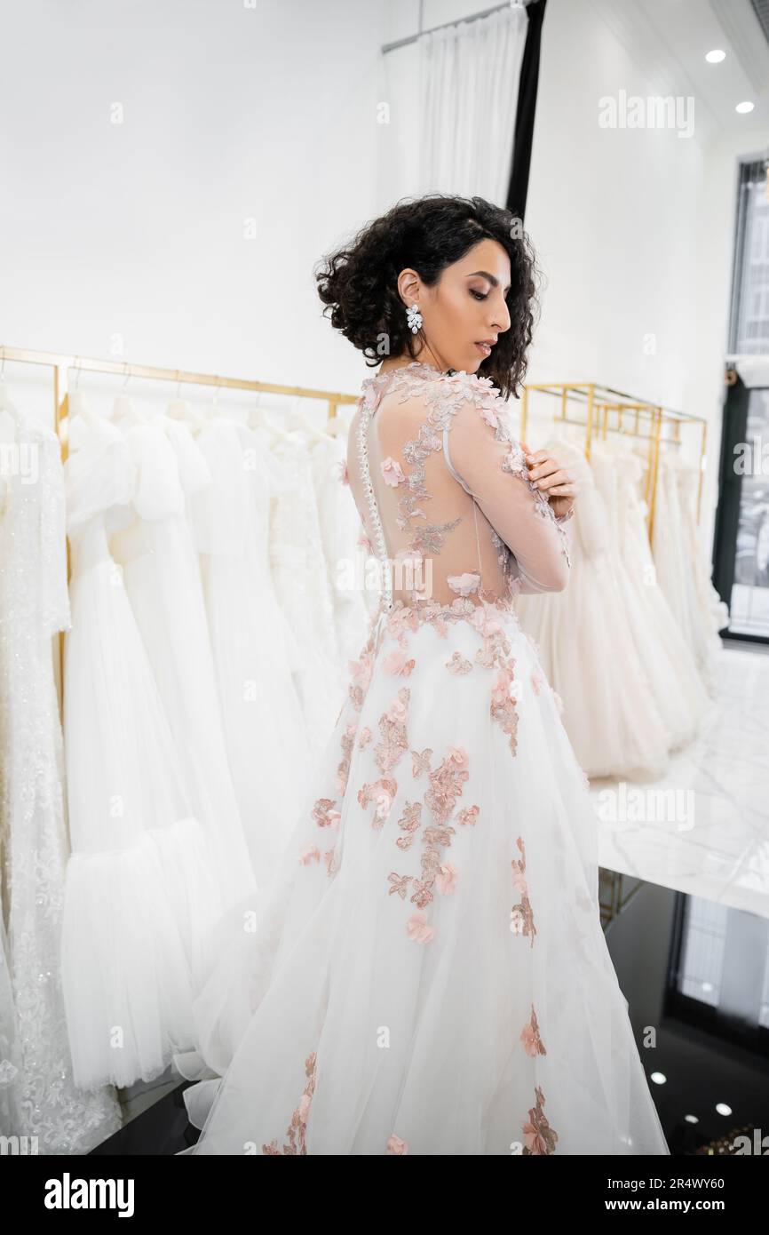 elegant middle eastern and brunette woman with wavy hair standing in gorgeous and floral wedding dress inside of luxurious bridal salon around white t Stock Photo