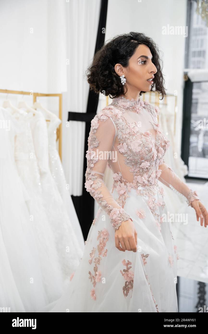 bride-to-be, surprised middle eastern and brunette woman with wavy hair standing in floral wedding dress inside of luxurious bridal salon around white Stock Photo