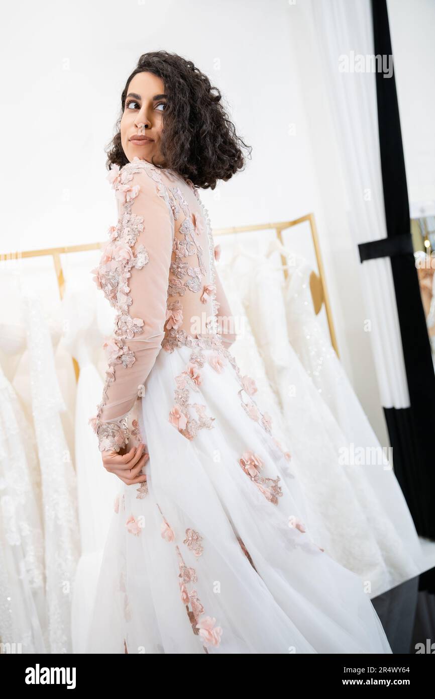 delightful middle eastern woman with wavy hair standing in gorgeous and floral wedding dress and looking away inside of luxurious salon around white t Stock Photo