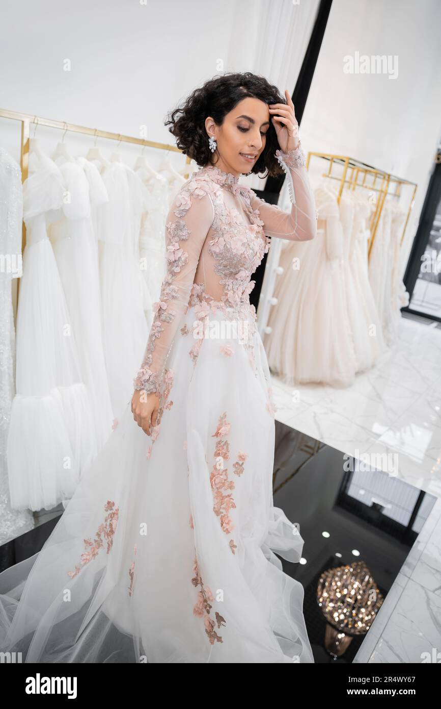 bride-to-be, charming middle eastern and brunette woman with wavy hair standing in gorgeous and floral wedding dress inside of luxurious bridal salon Stock Photo