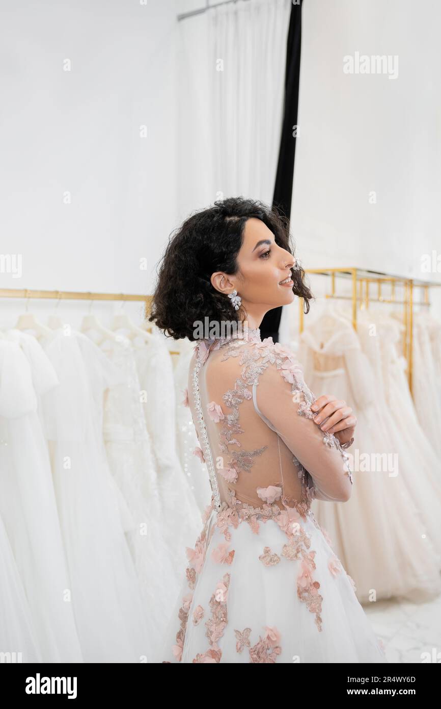 stunning middle eastern and brunette woman with wavy hair standing in gorgeous and floral wedding dress inside of luxurious bridal salon around white Stock Photo