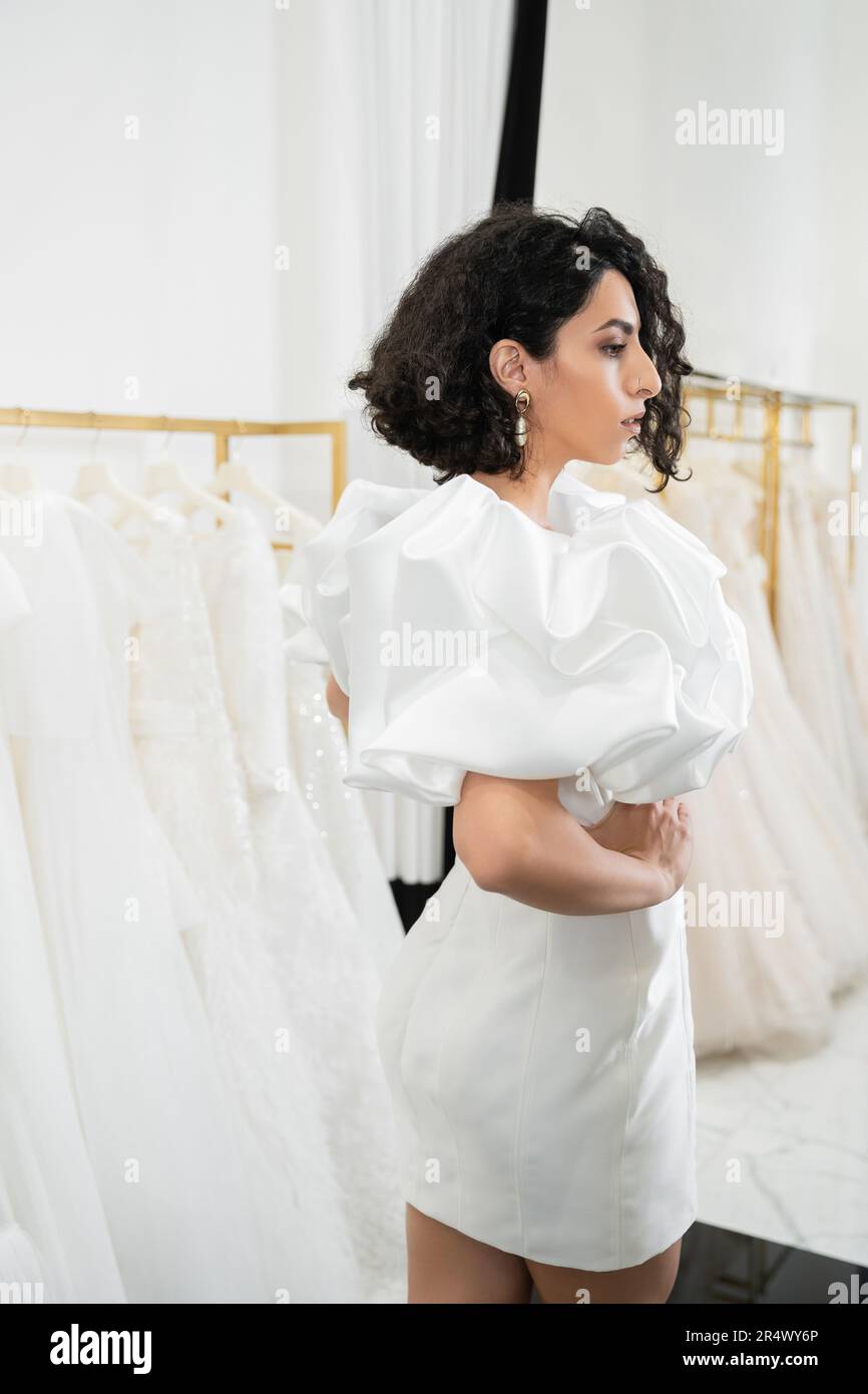 brunette middle eastern bride with brunette and wavy hair posing in trendy wedding dress with puff sleeves and ruffles in bridal boutique next to tull Stock Photo