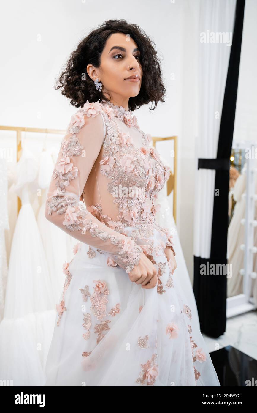 captivating middle eastern woman with wavy hair standing in floral wedding dress and looking away inside of luxurious salon around white tulle fabrics Stock Photo