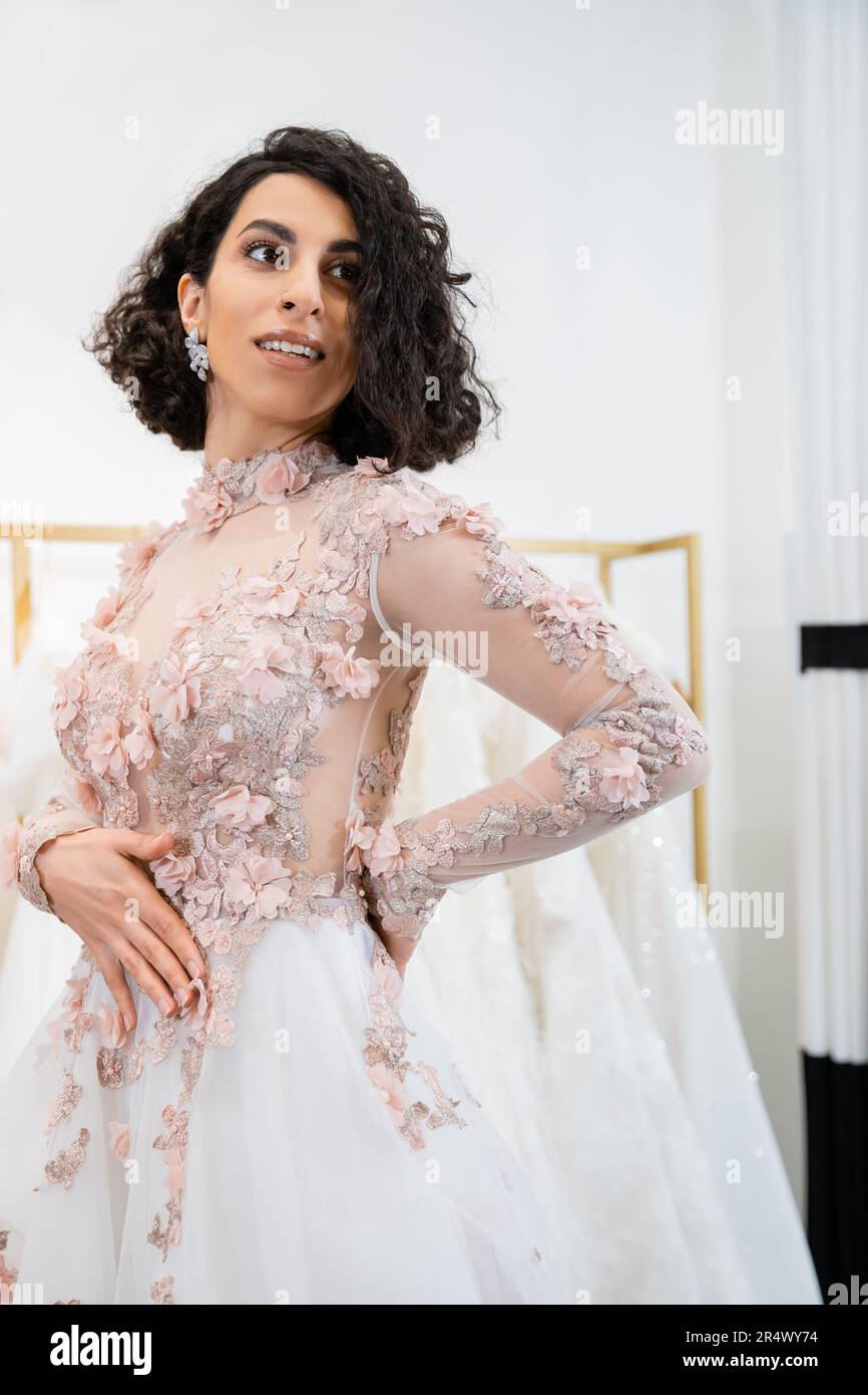 cheerful middle eastern and brunette woman with wavy hair standing in elegant and floral wedding dress and looking away inside of luxurious salon arou Stock Photo