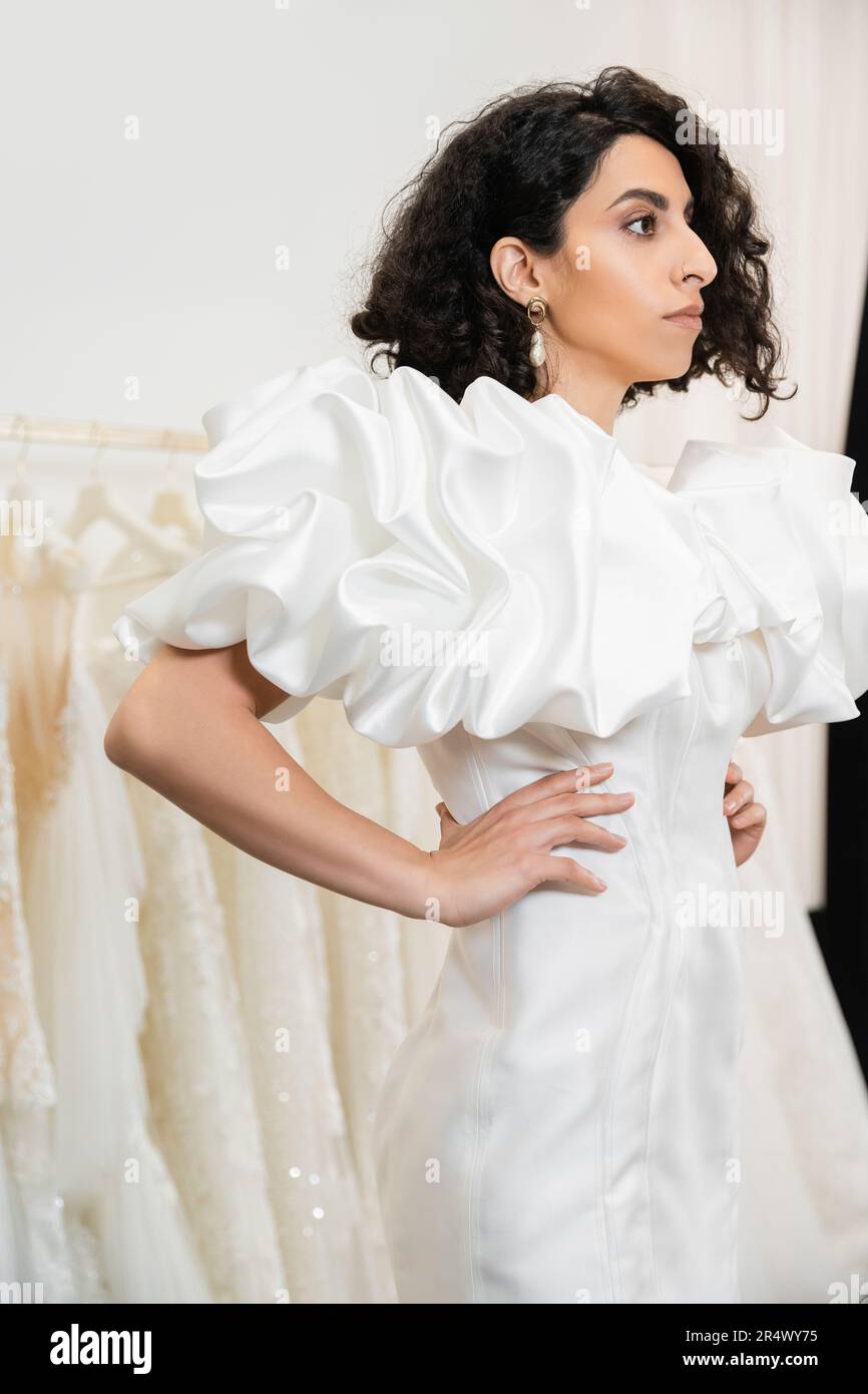 brunette middle eastern bride with brunette and wavy hair posing with hands on hips in trendy wedding dress with puff sleeves and ruffles in bridal sa Stock Photo