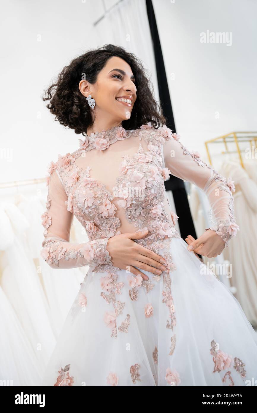 enchanting middle eastern and brunette woman with wavy hair standing in stunning and floral wedding dress and looking away inside of luxurious salon a Stock Photo