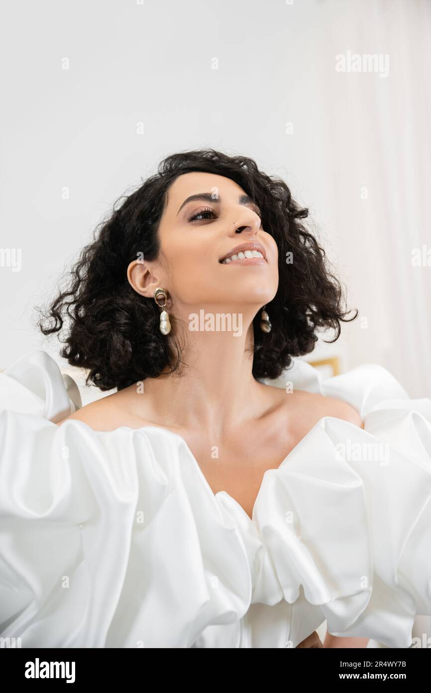 portrait of happy middle eastern bride with brunette and wavy hair posing in trendy wedding dress with puff sleeves and ruffles looking up in bridal s Stock Photo