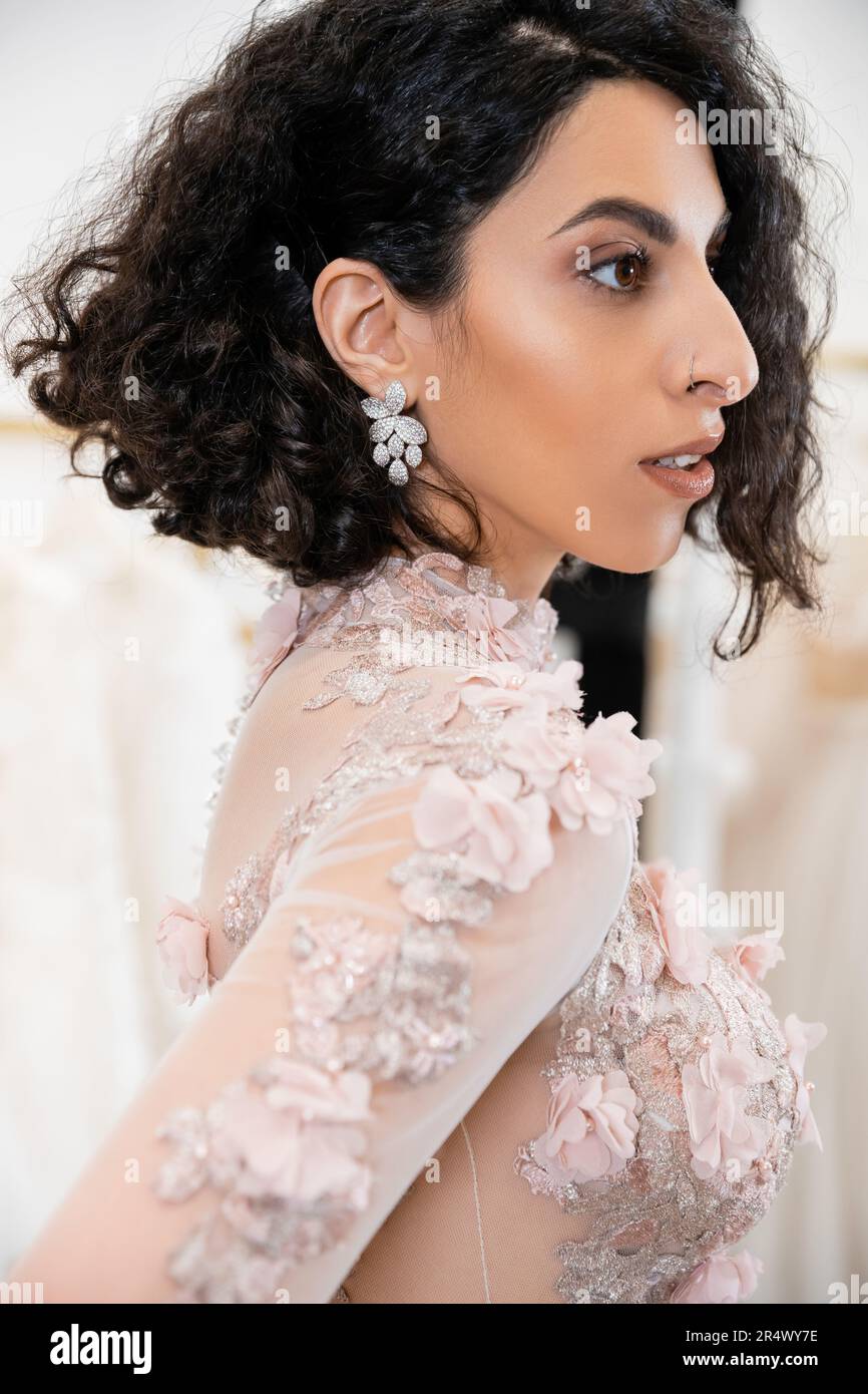 portrait of delightful middle eastern woman with wavy hair standing in gorgeous and floral wedding dress and looking away inside of luxurious salon ar Stock Photo