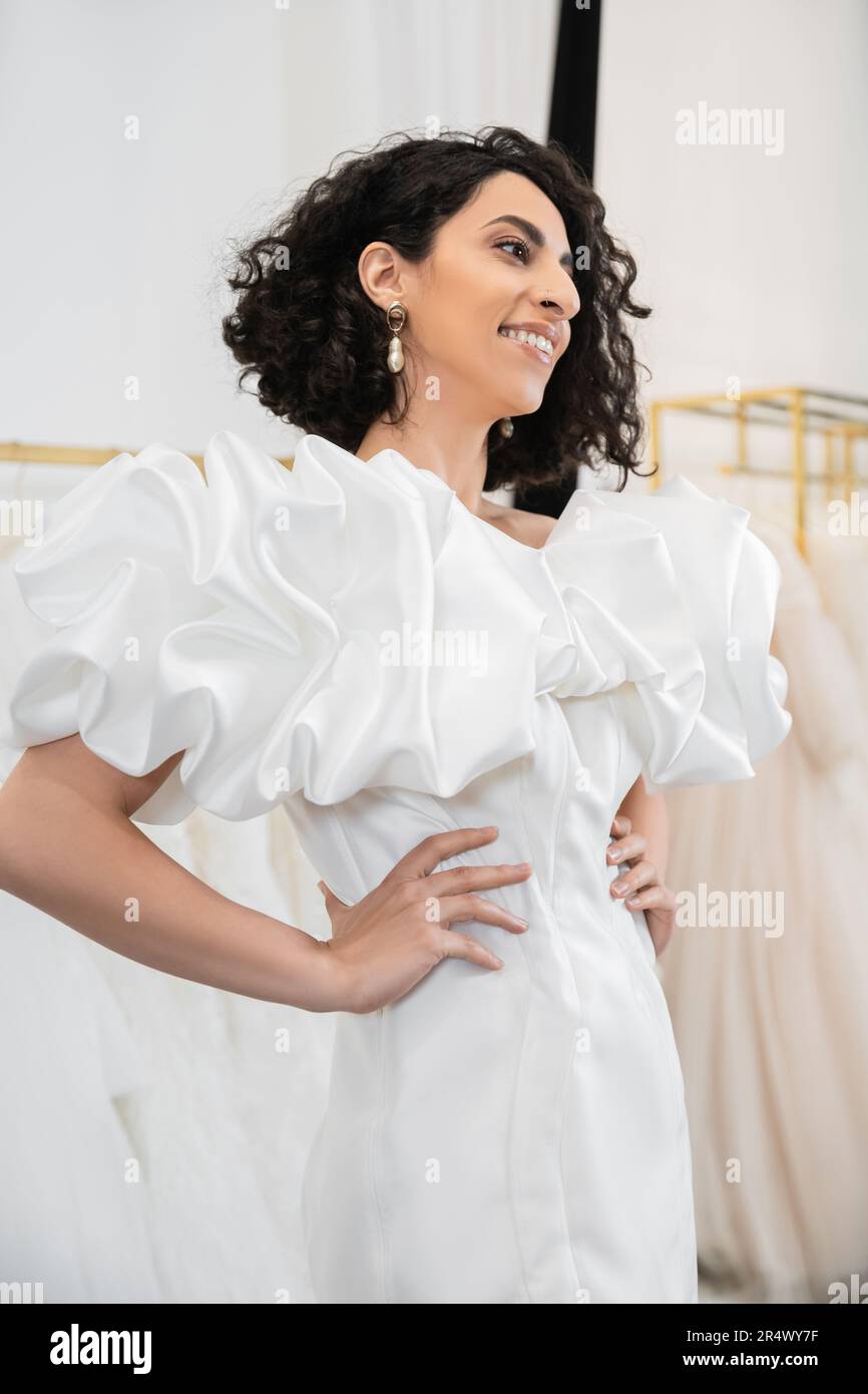 cheerful middle eastern bride with brunette and wavy hair posing with hands on hips in trendy wedding dress with puff sleeves and ruffles in bridal sa Stock Photo
