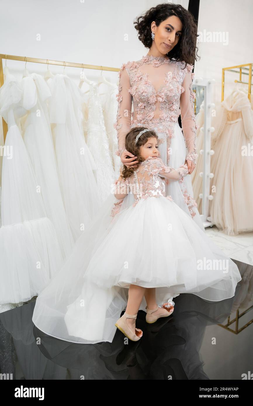 cute girl in floral attire hugging elegant woman with brunette hair standing in wedding dress near blurred white gown inside of luxurious bridal salon Stock Photo