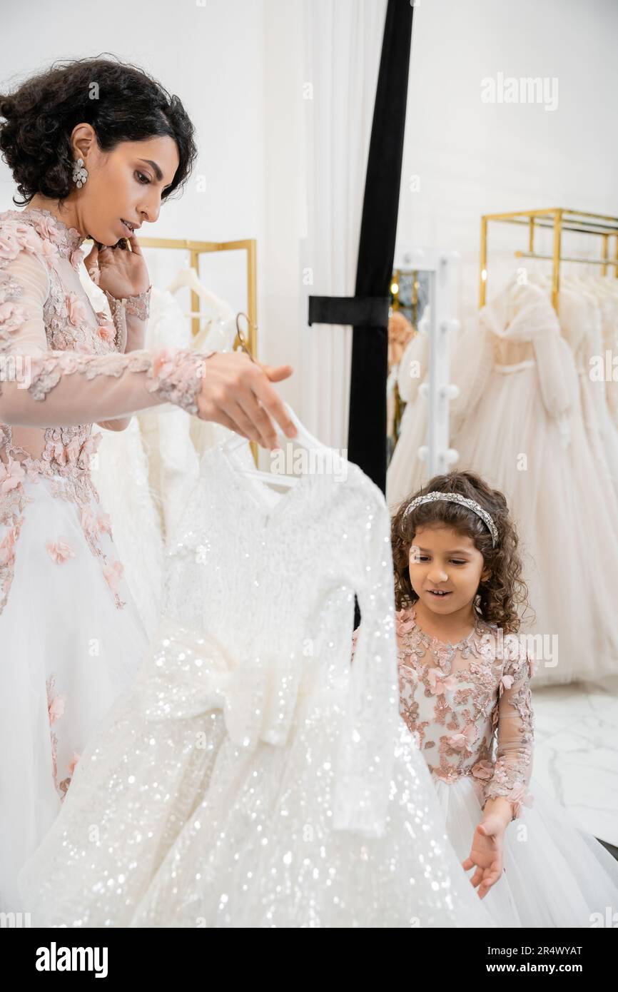 brunette middle eastern bride in floral wedding gown helping to choose dress for her little daughter in bridal salon around white tulle fabrics, proce Stock Photo