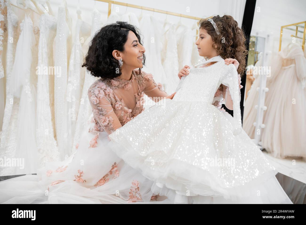 happy and brunette middle eastern woman in floral wedding gown helping to choose white dress for her little daughter in bridal salon around tulle fabr Stock Photo