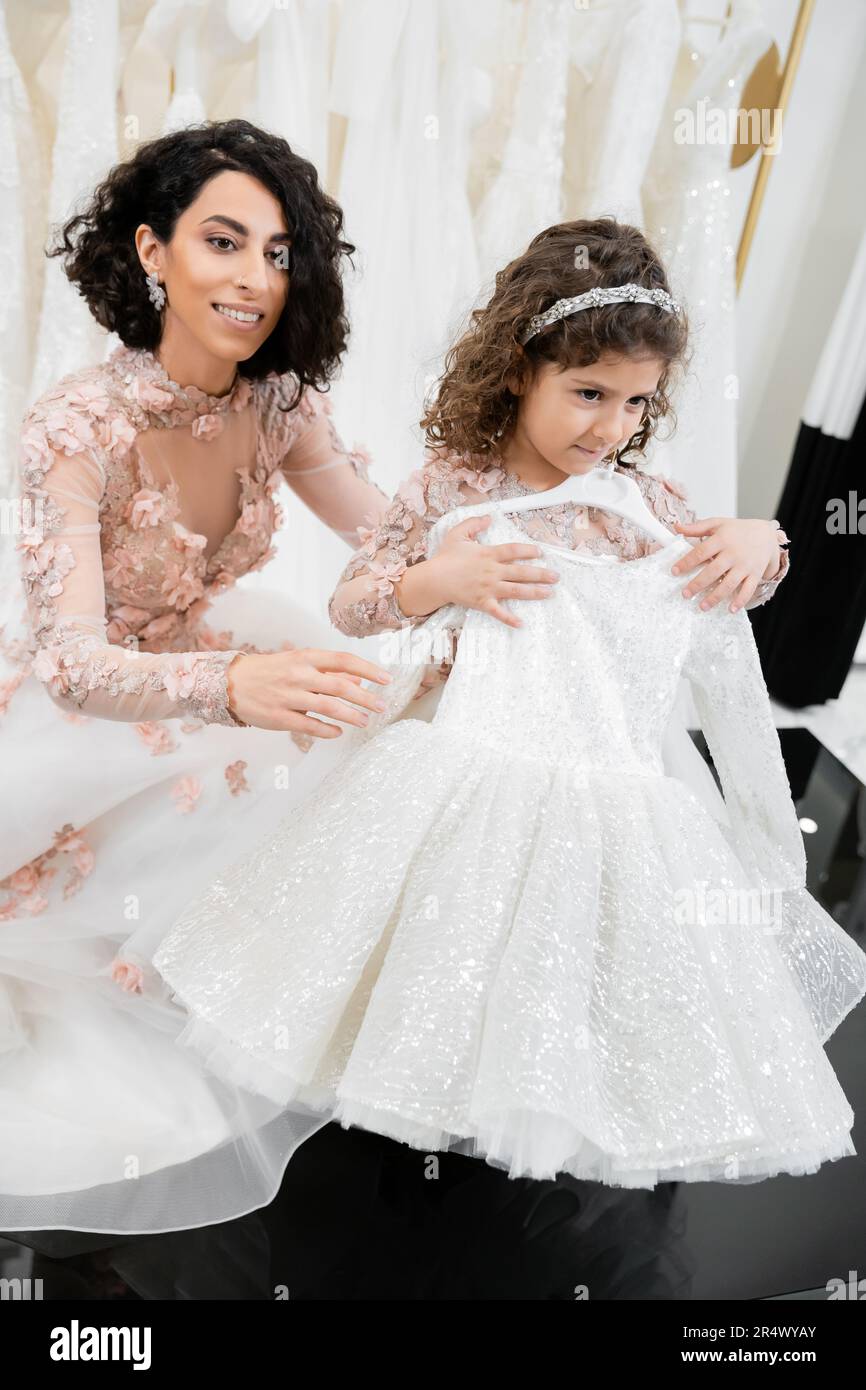 happy and brunette middle eastern bride in floral wedding gown helping to choose dress for her little daughter in bridal salon around white tulle fabr Stock Photo