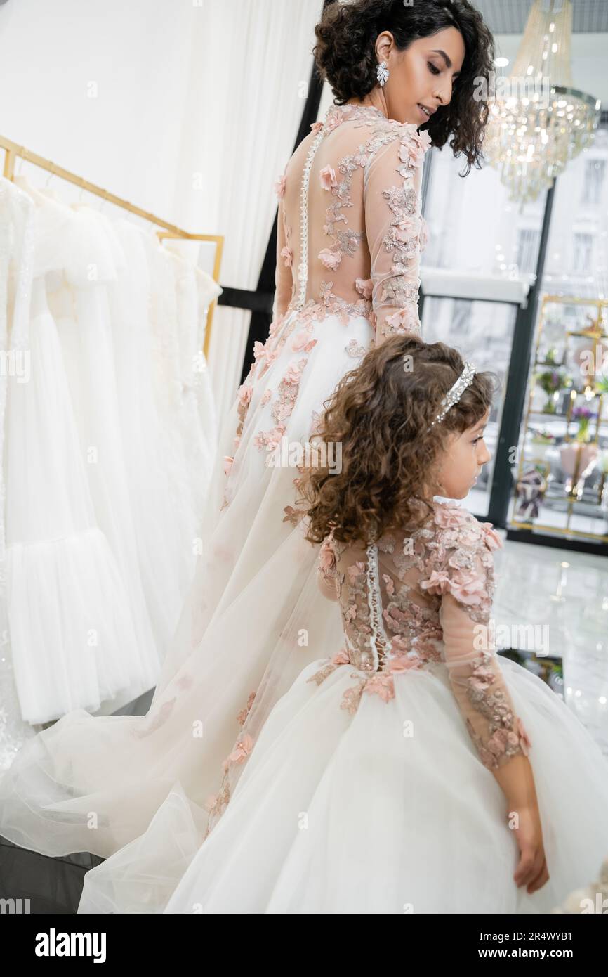 cute middle eastern girl in floral attire holding hands with woman and walking in floral dresses near blurred wedding gown inside of bridal salon, sho Stock Photo