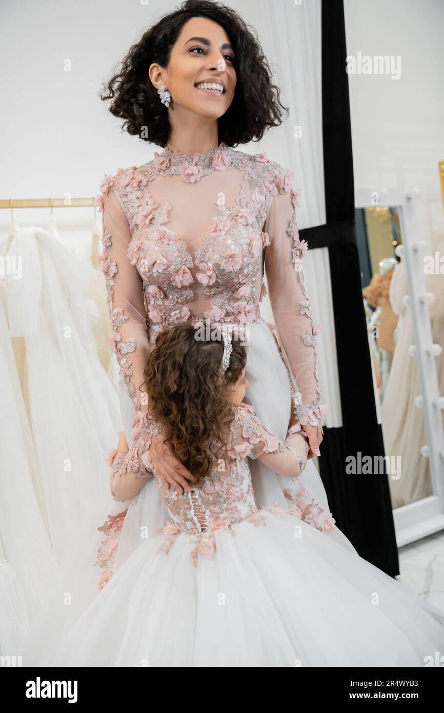 cute girl in floral attire hugging cheerful mother with brunette hair standing in wedding dress near blurred white gown inside of luxurious bridal sal Stock Photo