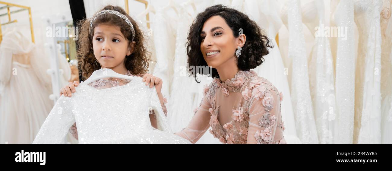 enchanting and brunette middle eastern bride in floral wedding gown helping to choose dress for her little daughter in bridal salon around white tulle Stock Photo