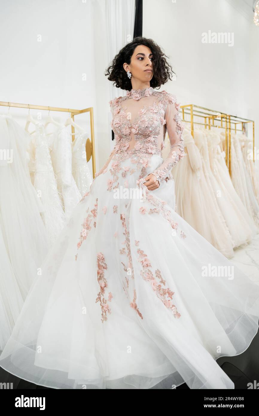 delightful and middle eastern woman with wavy hair trying on elegant and floral wedding dress inside of luxurious bridal salon, shopping, bride-to-be, Stock Photo