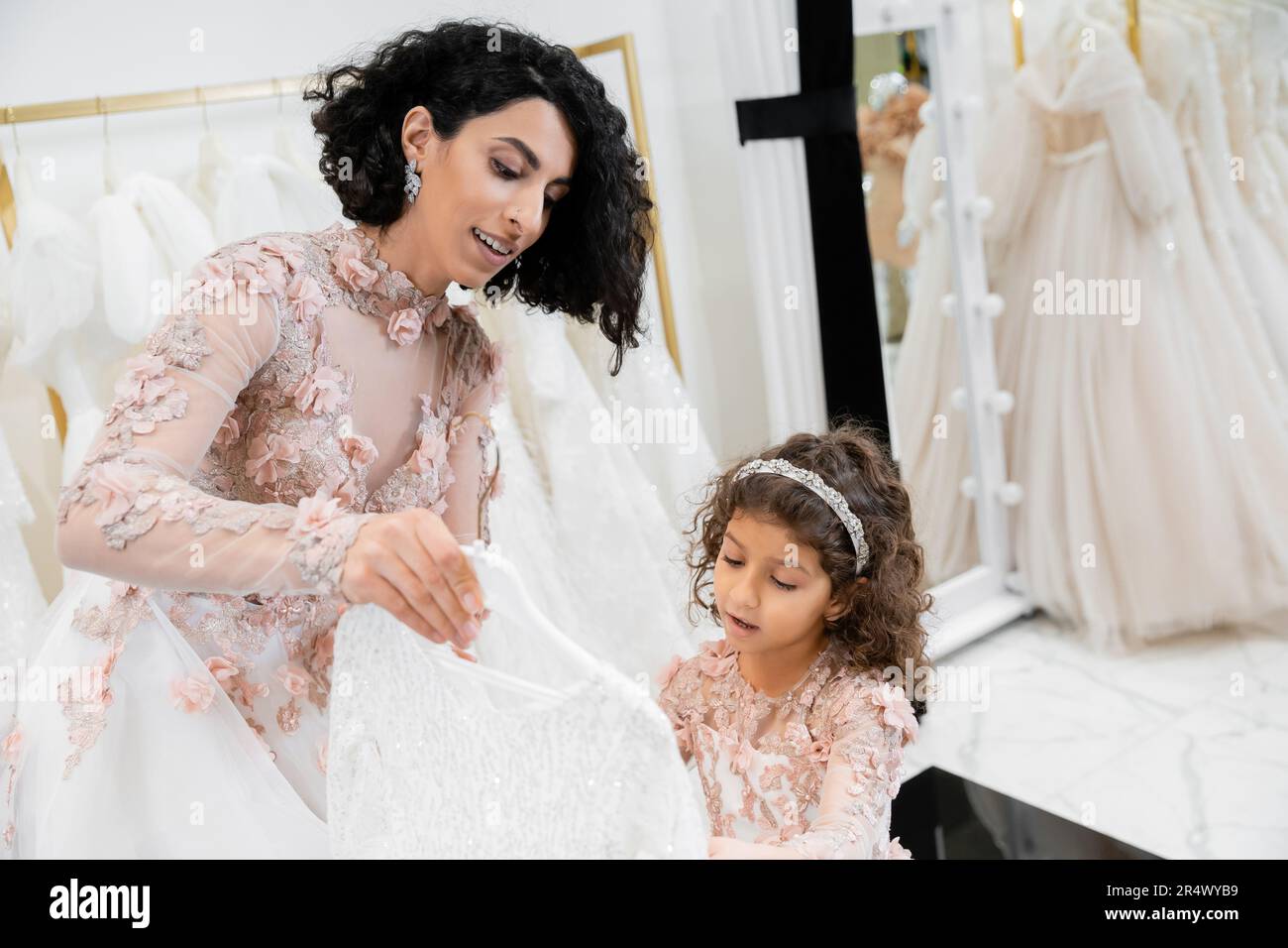 brunette middle eastern bride in floral wedding gown helping to choose dress for her cute little daughter in bridal boutique around white tulle fabric Stock Photo