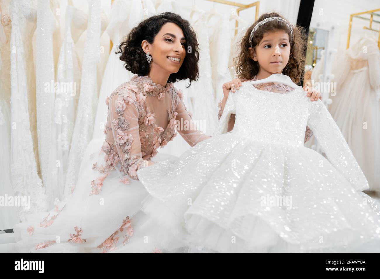 cheerful and brunette middle eastern bride in floral wedding gown looking away near her little daughter with cute dress in bridal salon around white t Stock Photo