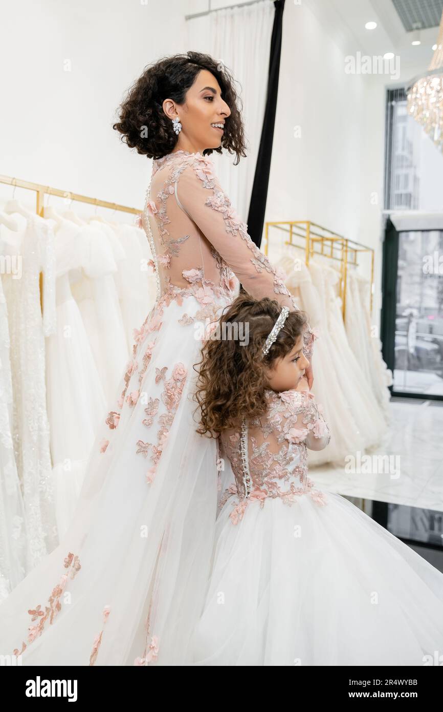 cute middle eastern kid in floral attire holding hands with happy woman and walking in floral dresses near blurred wedding gown inside of bridal salon Stock Photo