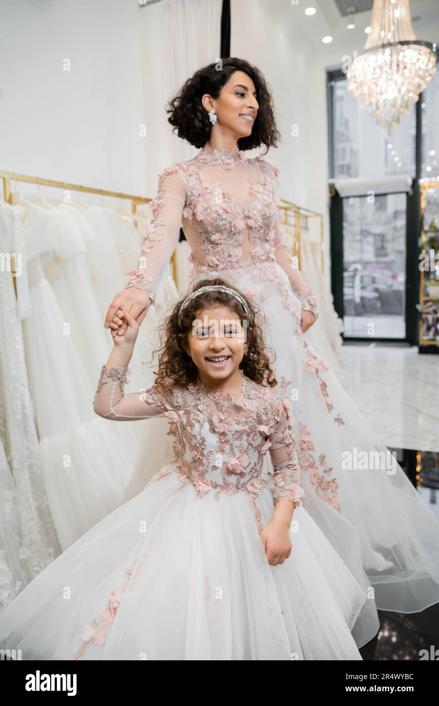 middle eastern girl in floral attire holding hands with cheerful woman standing in wedding dress near blurred white gown inside of luxurious bridal sa Stock Photo