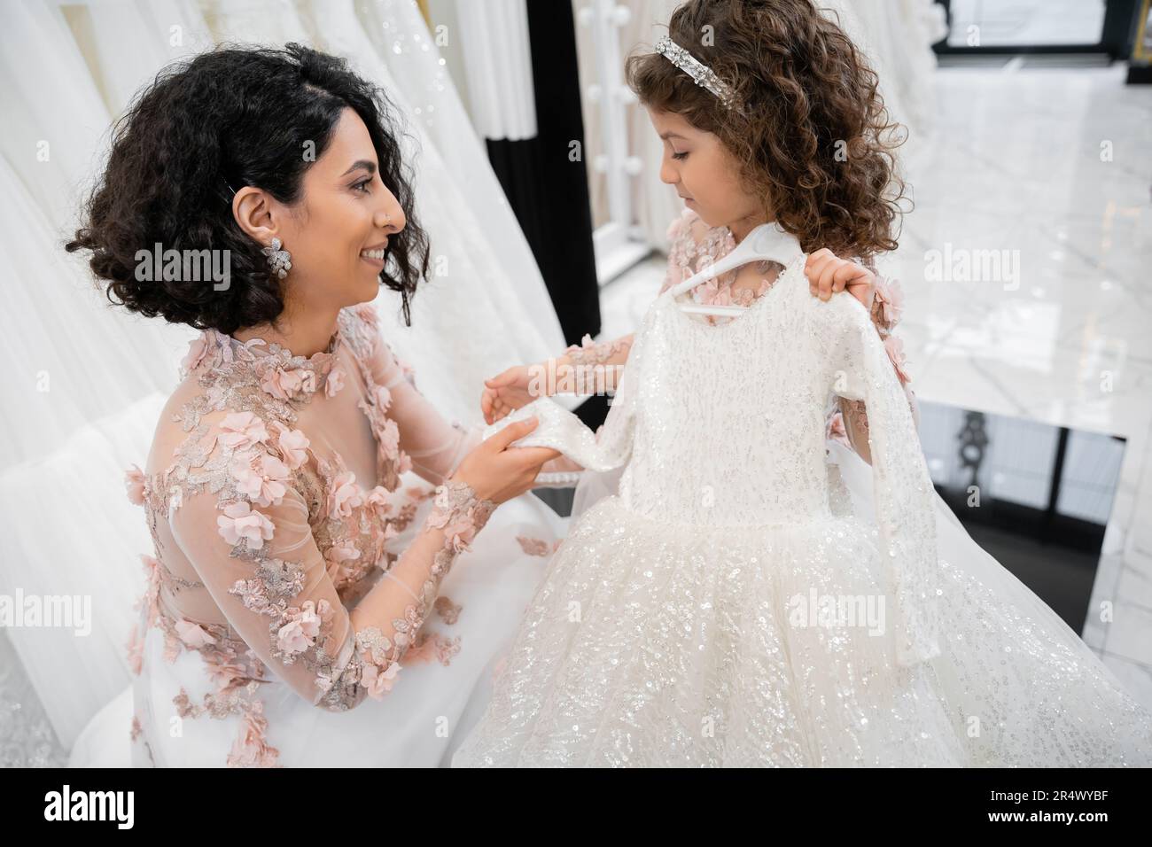 happy and brunette middle eastern bride in floral gown helping her little daughter to choose dress in bridal salon around white tulle fabrics, process Stock Photo