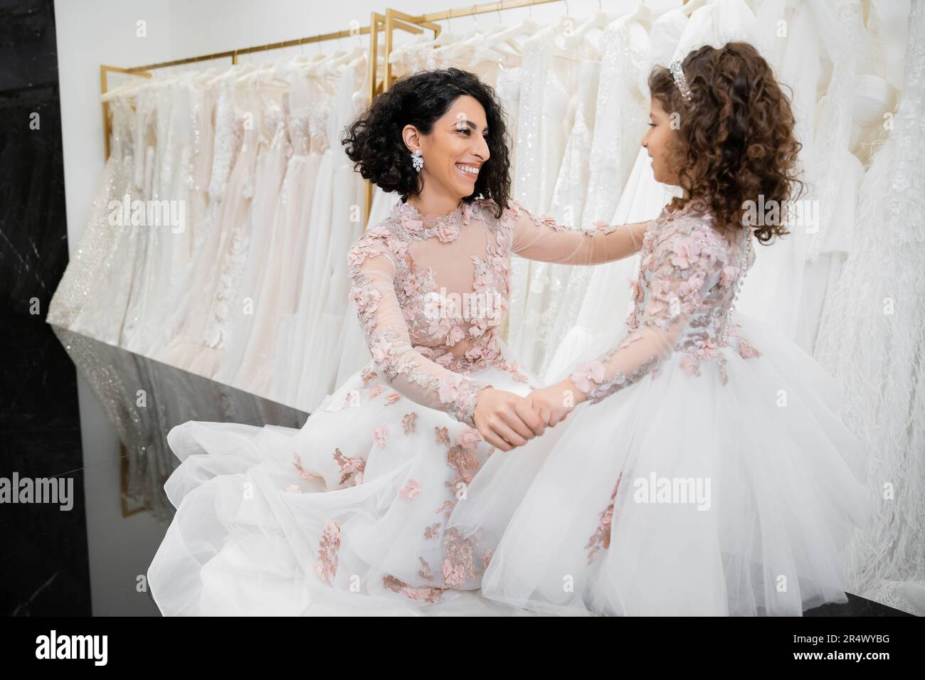 special moment, happy middle eastern bride in wedding gown sitting and holding hands with her little daughter in bridal salon around white tulle fabri Stock Photo