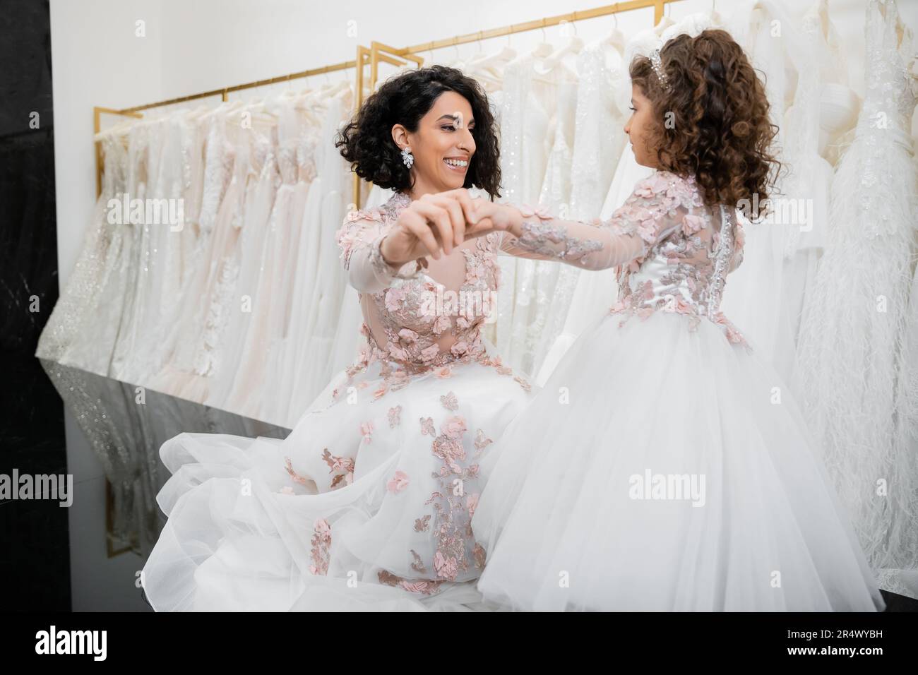 special moment, happy middle eastern bride in floral wedding gown sitting and holding hands with her little daughter in bridal salon around white tull Stock Photo