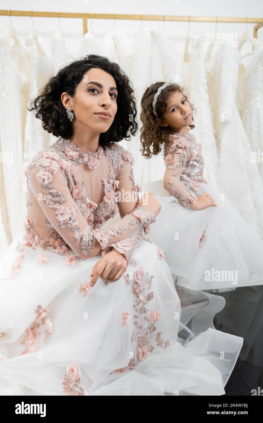 special moment, brunette middle eastern bride in floral wedding gown sitting next to her little daughter in bridal salon around white tulle fabrics, b Stock Photo