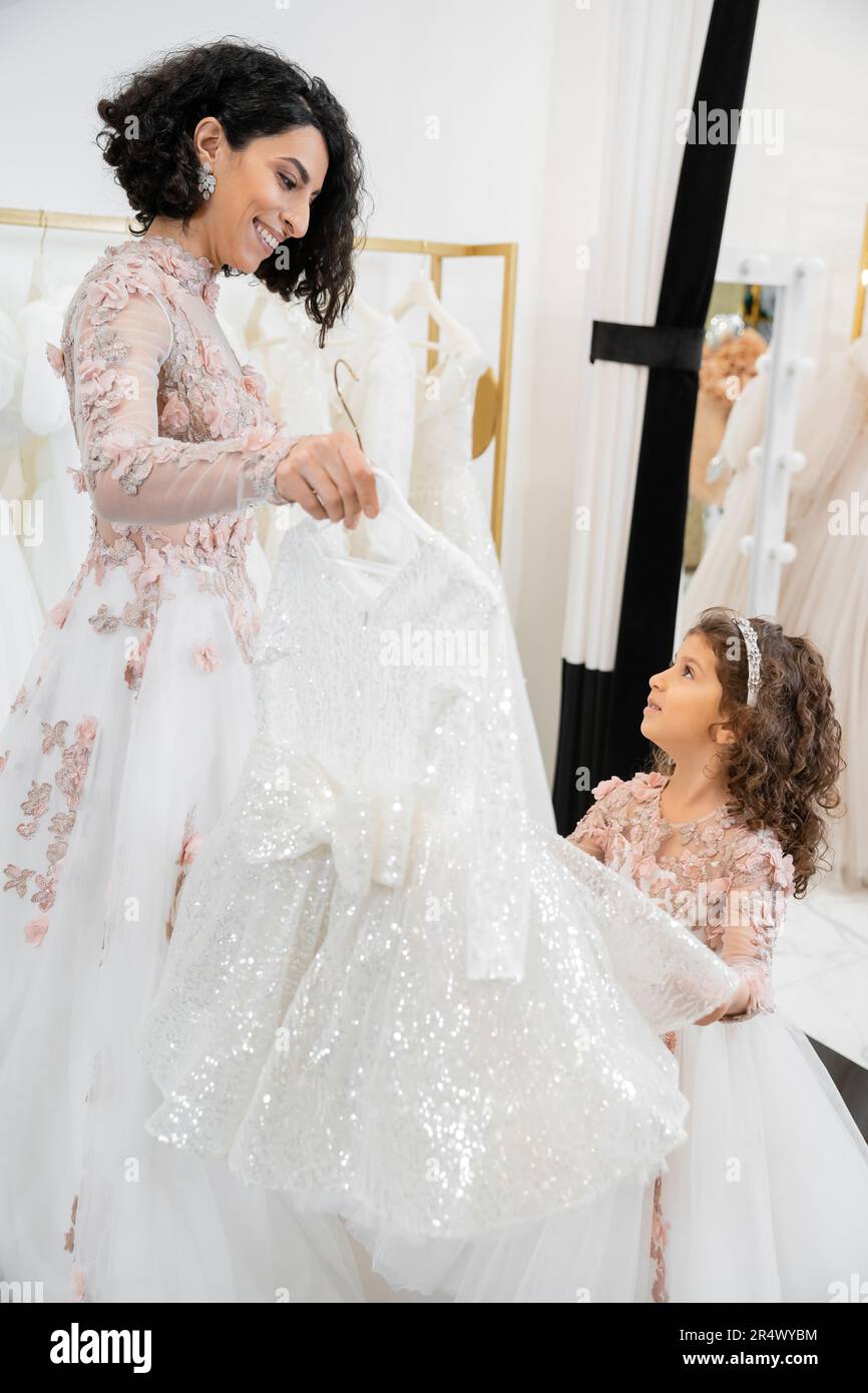 happy and brunette middle eastern bride in floral wedding gown helping to choose dress for her little daughter in bridal salon around white tulle fabr Stock Photo