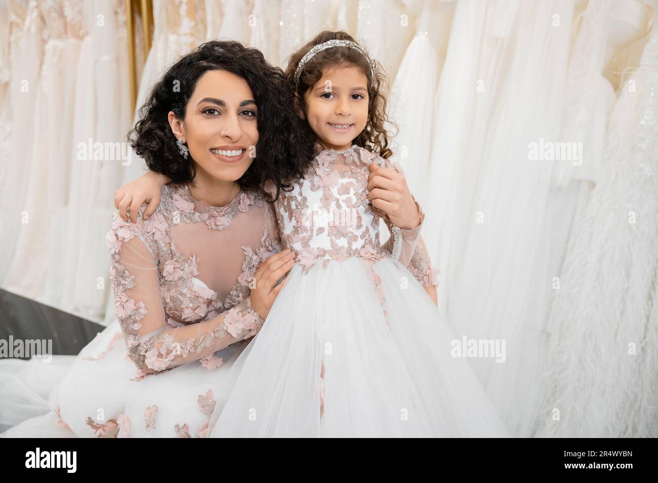 special moment, happy middle eastern woman in floral wedding gown sitting and embracing her little daughter in bridal salon around white tulle fabrics Stock Photo