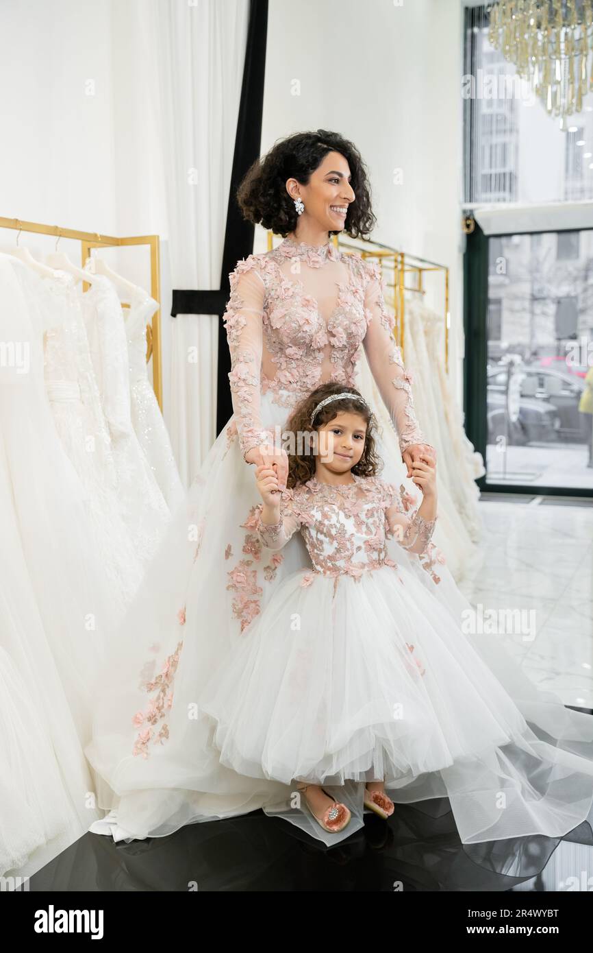 cute middle eastern girl in floral attire holding hands with cheerful woman standing in wedding dress near blurred white gown inside of bridal salon, Stock Photo