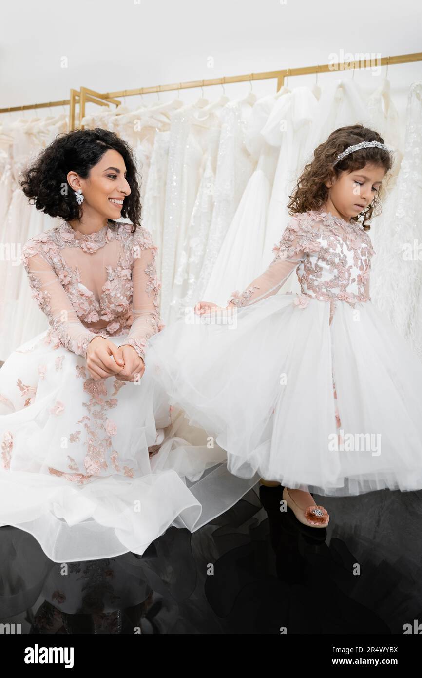 special moment, joyful middle eastern bride in floral wedding gown sitting and looking at little daughter in bridal salon posing around white tulle fa Stock Photo