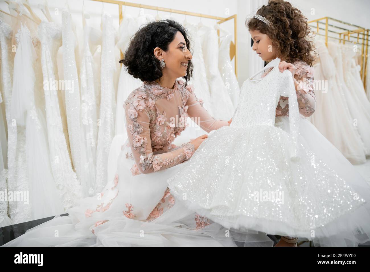 blissful and brunette middle eastern bride in floral wedding gown helping to choose dress for her cute little daughter in bridal salon around white tu Stock Photo