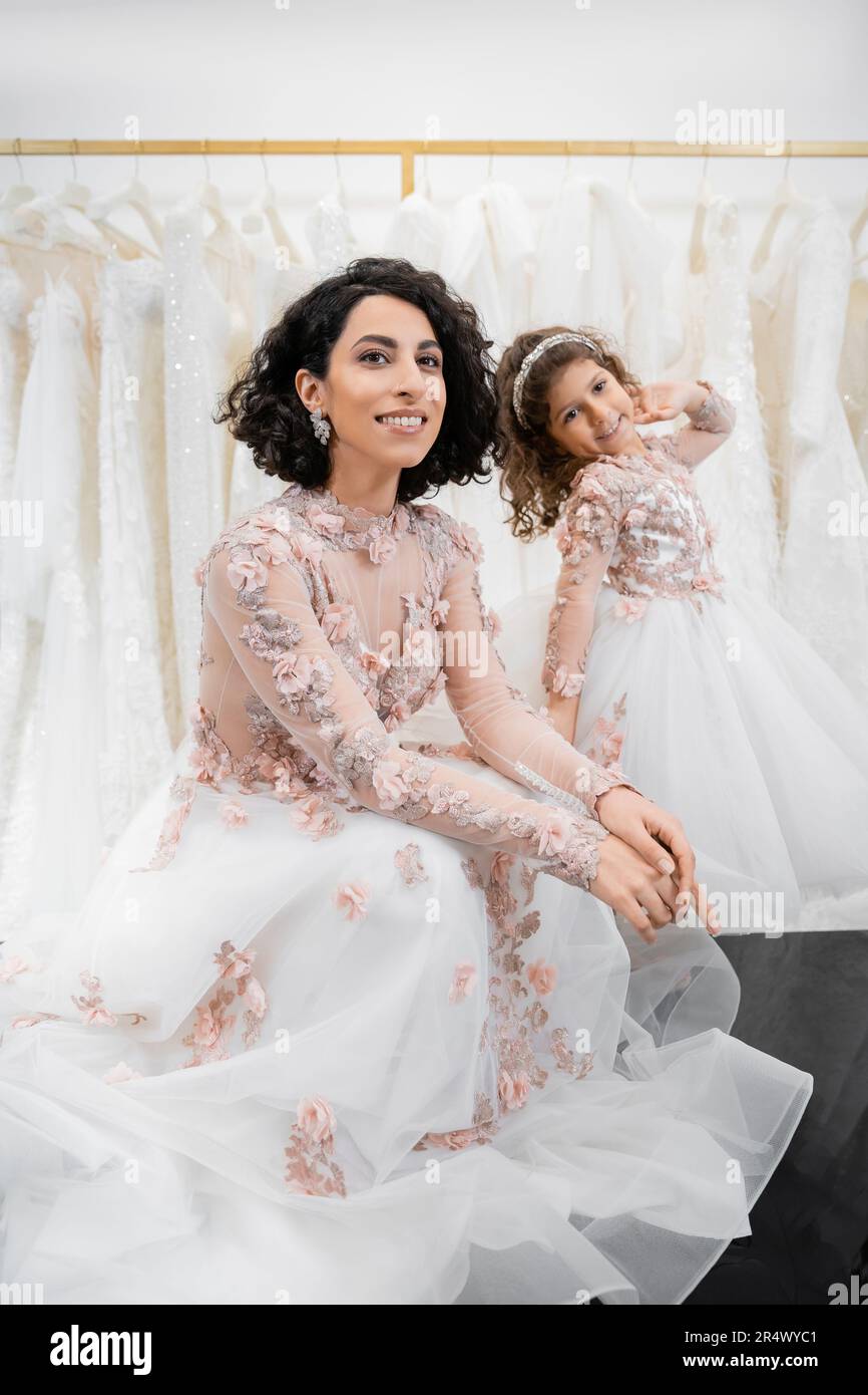 special moment, charming and happy middle eastern bride in floral wedding gown sitting next to her little daughter in bridal salon around white tulle Stock Photo